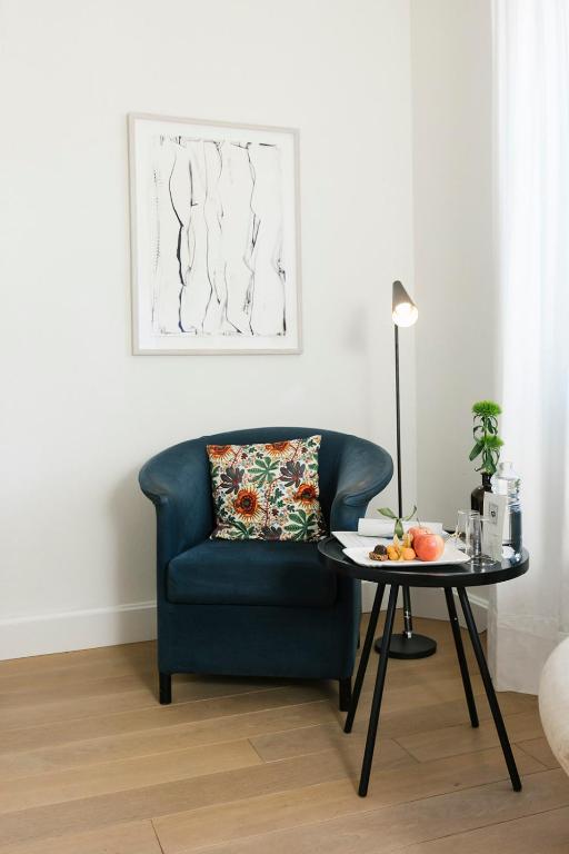 Mūsdienīgs stūrītis ar zilu krēslu un spilvenu ar ziedu motīvu, blakus melnā galdiņa ar uzkodām un lampu, kas piešķir siltumu telpai.