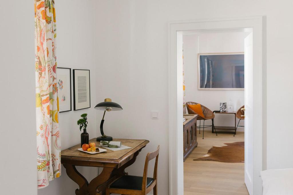 Interjera skats ar koka galdu, lampu un dekoratīvām aizkariem, kas vijas pie loga. Aiz durvīm redzama moderna telpa ar dīvānu un mākslas darbiem uz sienām.