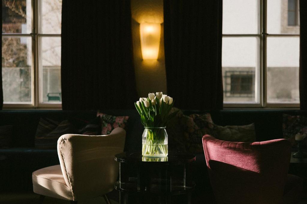 Gaiša un eleganta kafijas istaba ar ziedu pušķi stikla vāzē un komfortablām krēsliem. Dabas gaisma plūst caur logiem, radot mājīgu atmosfēru.