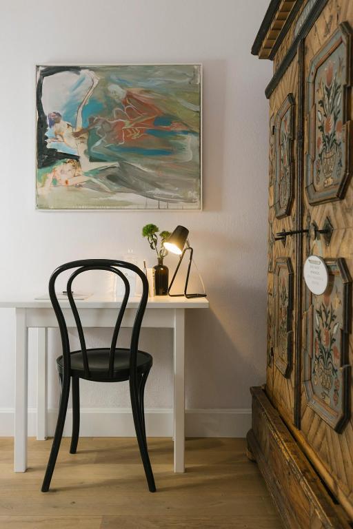 Mūsdienīgs rakstāmgalds ar melnu krēslu un lampu, blakus stilizētam mākslas darbam un greznu koka skapīti.