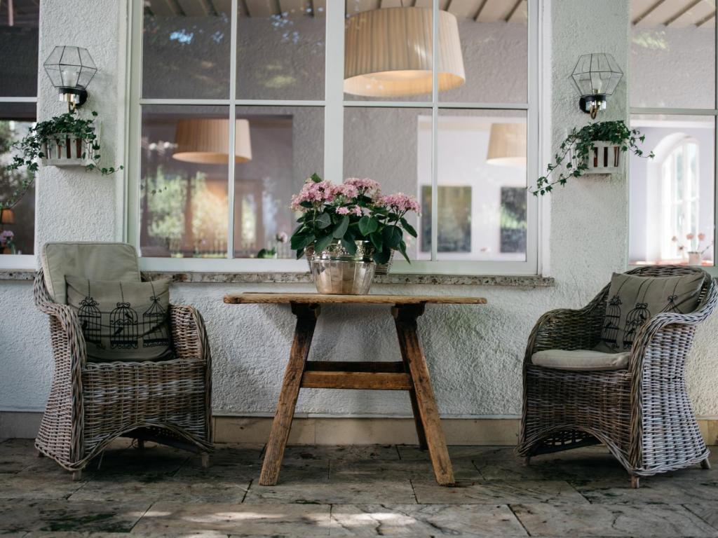 Elegantā terases zona ar divām ratiņu krēslām un koka galdu, uz kura novietota ziedu kompozīcija, radot siltu un mājīgu atmosfēru.