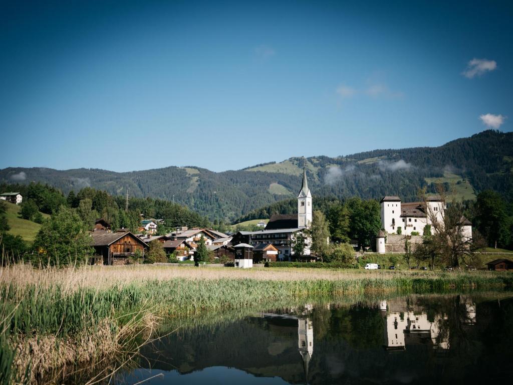 Pilsētiņa kalnu apkārtnē ar gleznainiem mājokļiem un baznīcu, kas atspoguļojas ūdenī. Skats uz kalniem un zaļajām pļavām.