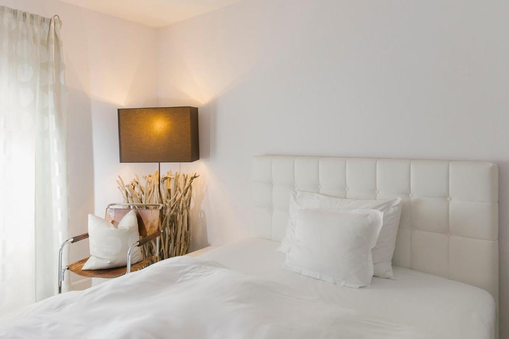 Mūsdienīga guļamistaba ar baltu gultu, spilveniem un stilīgu lampu, kas rada siltu atmosfēru. Interjers ir vienkāršs, ar dabīgām krāsām un vieglām aizkariem.