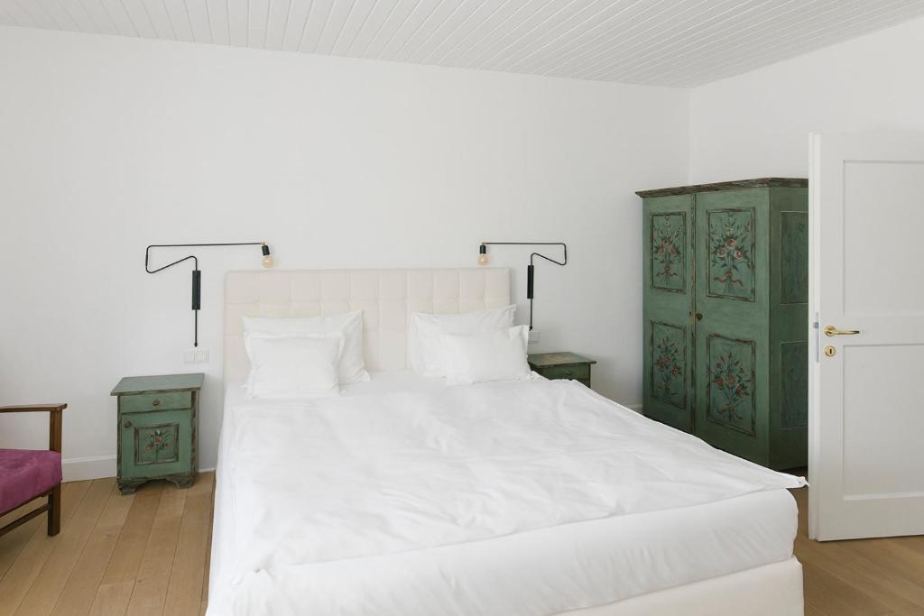 Mūsdienīga guļamistaba ar gultu un baltām gultasveļām, dekoratīvām lampām un senatnīgu zaļu skapīti. Telpa ir gaiša un eleganta, ar koka grīdu un minimālistisku dizainu.