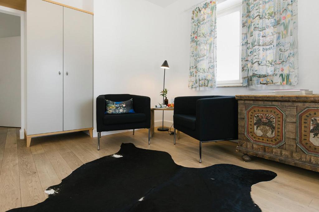 Mūsdienīgs dzīvojamās telpas interjers ar divām melnām krēslu sēdvietām, koka grīdu un dekoratīvu skapīti. Logi ir rotāti ar krāsainām aizkariem, radot mājīgu atmosfēru.