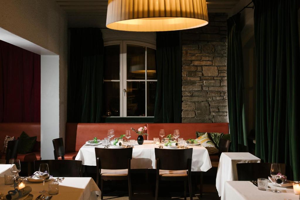 Elegants restorāna iekārtojums ar baltām galdautām un izsmalcinātu galda piederumu izvēli. Tumši zaļas aizkaras un akmens siena piešķir siltu atmosfēru.