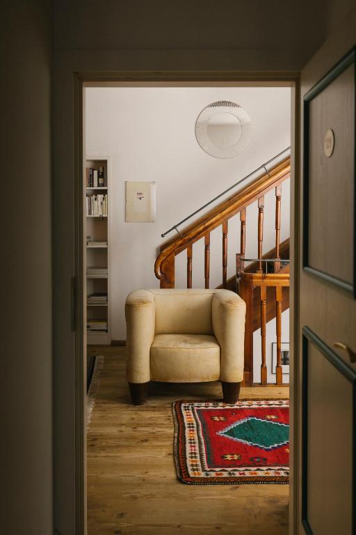 Mājīga telpa ar dīvānu un paklāju, kas vērsta uz koka kāpnēm. Gaisma ieplūst no durvīm, radot siltu atmosfēru.