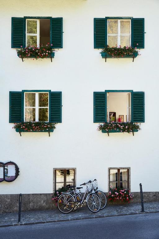 Eleganta mājas fasāde ar zaļām žalūzijām un puķu podiem pie logiem. Divas velosipēdu grupas novietotas pie mājas.