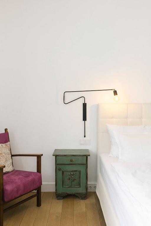 Mūsdienīgs guļamistabas interjers ar gultu, vintage stila naktsgaldiņu un stilīgu sienas lampu. Komfortabla krēsls ar rozā spilvenu papildina telpas estētiku.