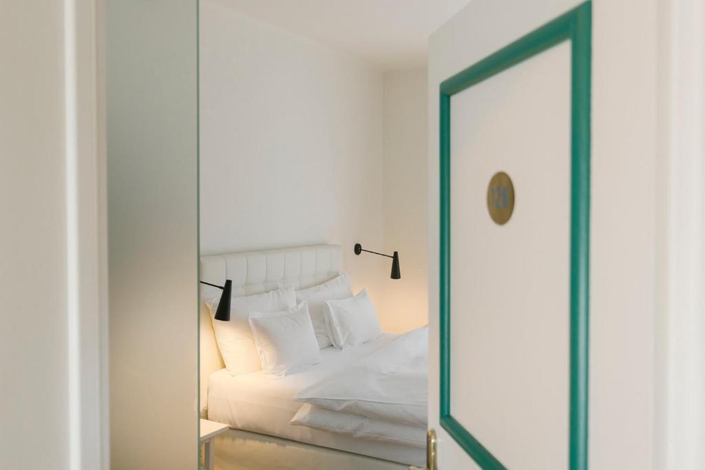 Mūsdienīga guļamistaba ar baltām gultas veļām un melnu gaismekli, redzama caur atvērtām durvīm. Guļamistaba ir gaiša un ērta, ar minimālistisku dizainu.