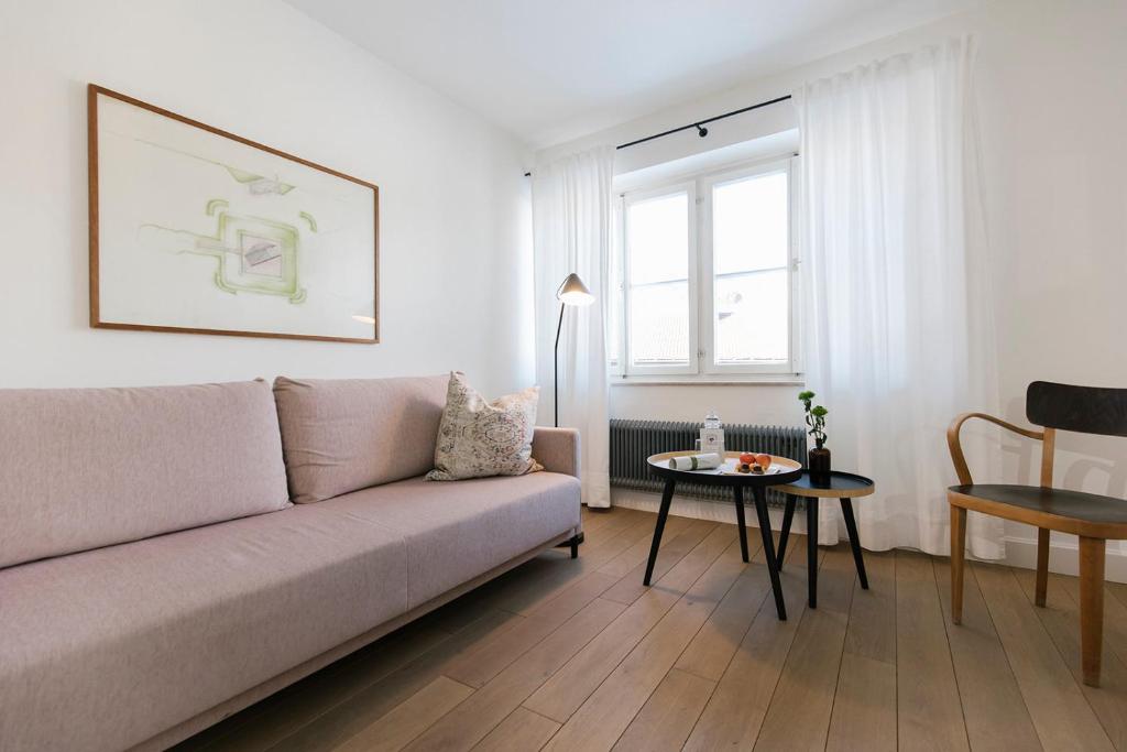 Mūsdienīgs viesistabas interjers ar gaišiem toņiem, rozā dīvānu un dekoratīvu spilvenu. Uz galda atrodas augļi un ūdens pudele, logi ar baltām aizkariem nodrošina dabīgu apgaismojumu.