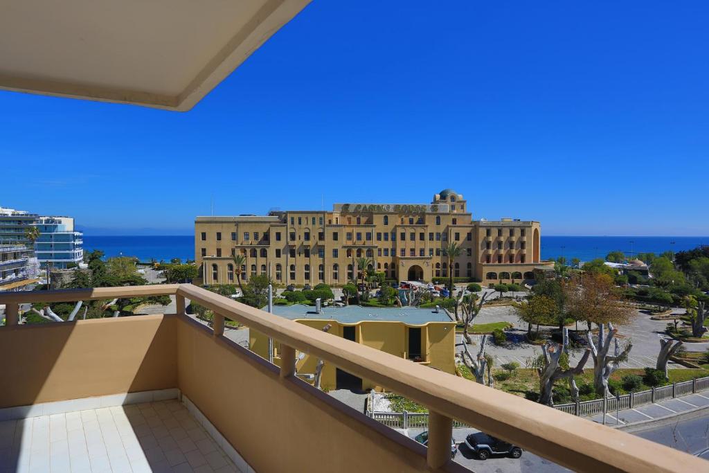 Skats no balkona uz piejūras viesnīcu ar zilo debesu un jūras fona. Šajā ainavā redzami dārzi un apzaļumojums, kas papildina viesnīcas arhitektūru.