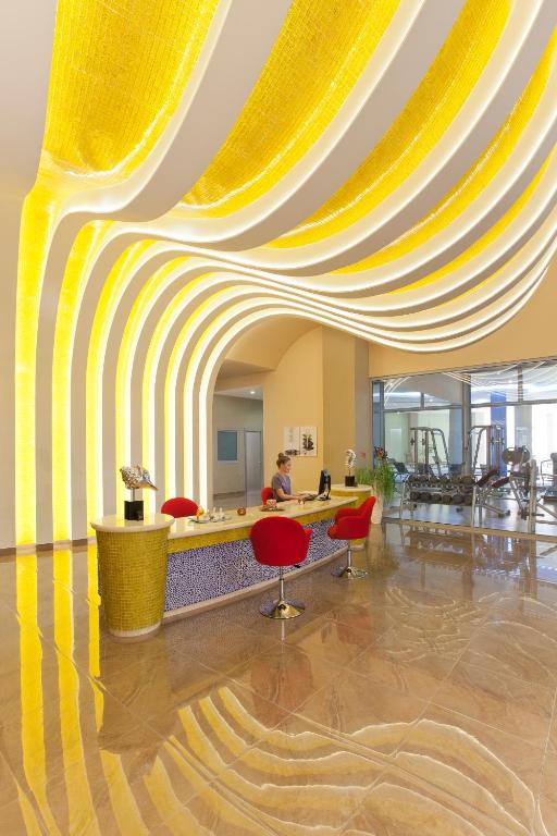 Mūsdienīga viesnīcas reģistratūra ar dzeltenu viļņotu sienu un stilīgām sarkanām krēsliem. Interjers ir gaišs un pievilcīgs, radot siltu un viesmīlīgu atmosfēru.