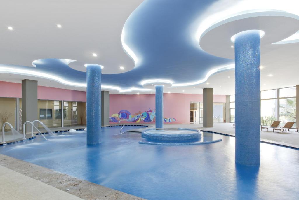 Mūsdienīgs iekštelpu baseins ar zilu ūdeni un stilīgiem gaismas elementiem, kas rada relaksējošu atmosfēru. Fona siena ar krāsainu gleznu piešķir telpai dzīvīgumu.