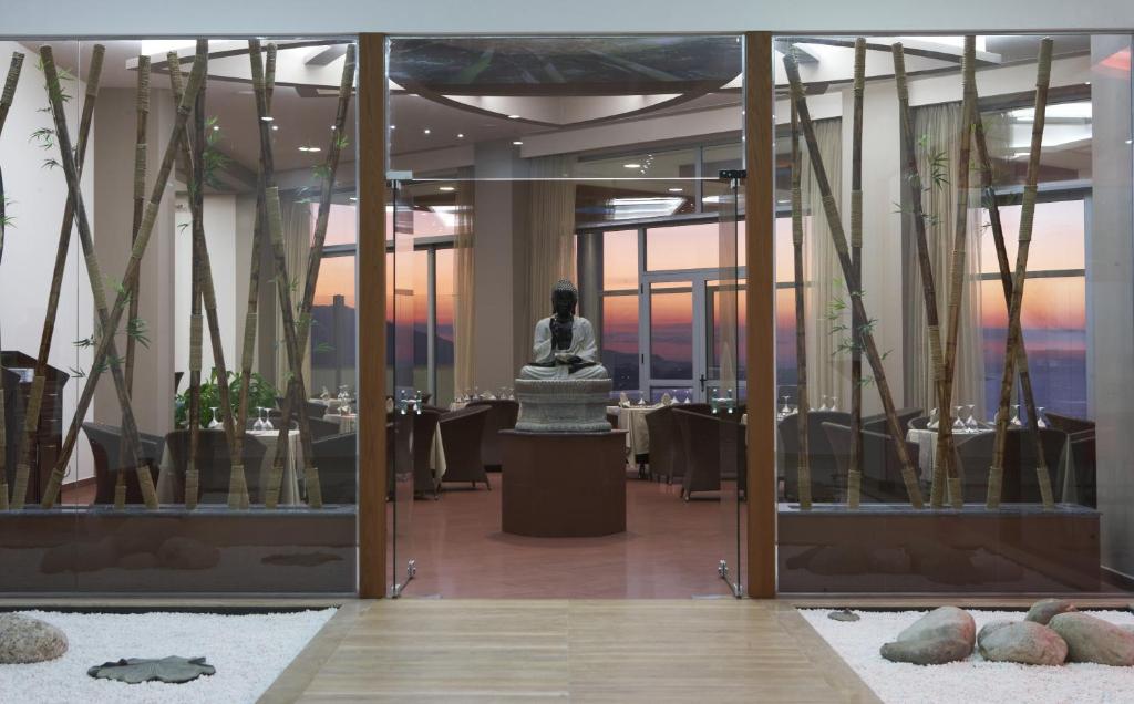 Mūsdienīga restorāna iekārtojums ar budas statuju centrā, apjoztu ar bambusa stabiem un skatu uz saulrietu caur lieliem logiem. Elegantās galdu klāšanas detaļas rada viesmīlīgu atmosfēru.