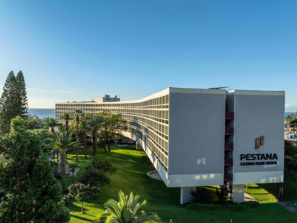 Pestana Casino Park Hotel ēka ar modernu arhitektūru, kas atrodas pie jūras, ieskauta tropiskā dārzā ar palmu kokiem.