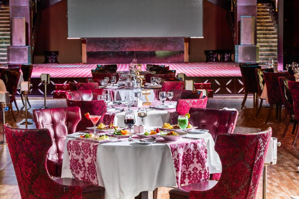 Elegants restorāns ar sarkaniem krēsliem un galdautiem, kas dekorēti ar izsmalcinātām rakstiem. Uz galdiem redzami dažādi dzērieni un ēdieni, bet fona skatuve gaida izrādēm.
