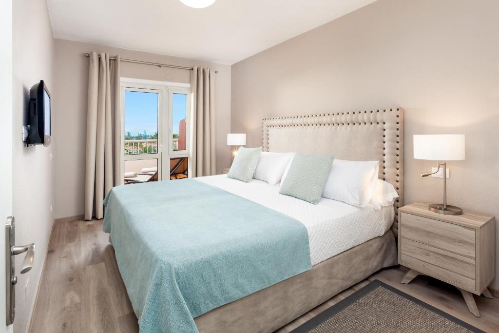 Moderns guļamistabas interjers ar ērtu divguļamo gultu, mīkstu segumu un lieliem logiem, caur kuriem redzams ainava. Sienas ir gaišas, un telpa iekārtota ar minimālistiskiem mēbeļu elementiem.