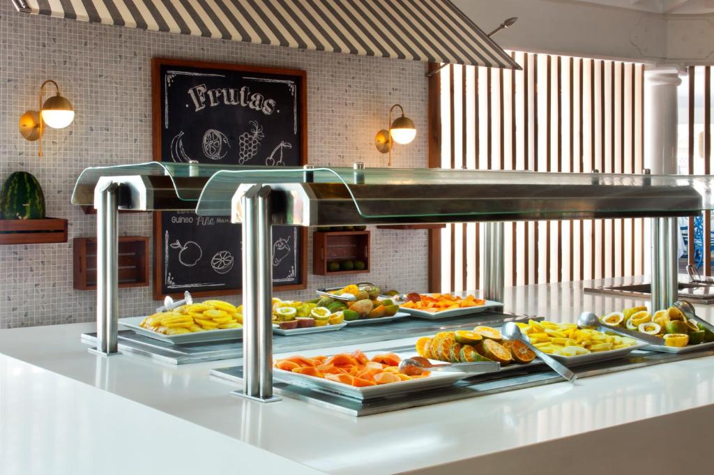 Svaigu augļu bufete ar dažādiem šķīvjiem un modernu dizainu, ideāli piemērota viesnīcu vai restorānu videi. Uz sienas redzama melnā tāfele ar augļu nosaukumiem.