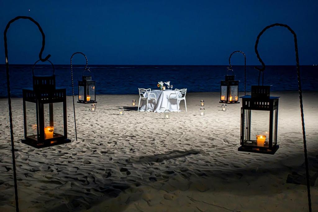 Romantiska vakara vakariņas uz pludmales, apgaismotas ar laternām un svecēm. Galda klājums ar ziediem un divām krēslām izveido intīmu atmosfēru.