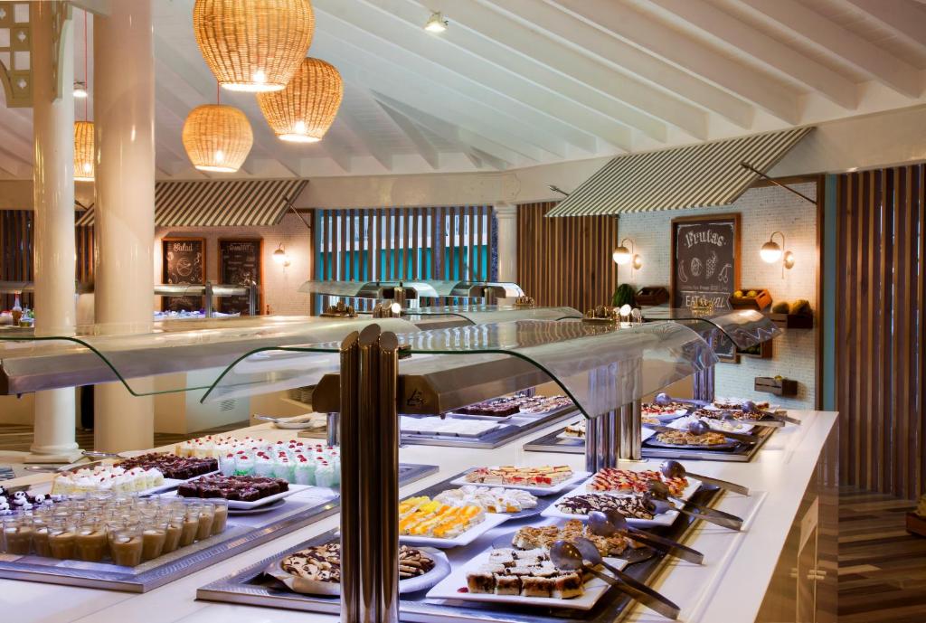 Bufete ar dažādiem saldumiem, kas izkārtoti uz galda ar stikla pārklājumu. Telpā ir gaiša apgaismojuma un dabīgu materiālu elementi, radot viesmīlīgu atmosfēru.