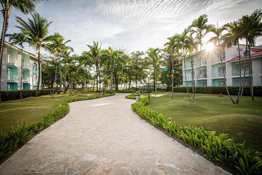 Skaists ceļš ved cauri zaļajai ainavai ar palmām, apņemot viesnīcas ēkas. Gaisma spīd starp kokiem, radot mierīgu atmosfēru.