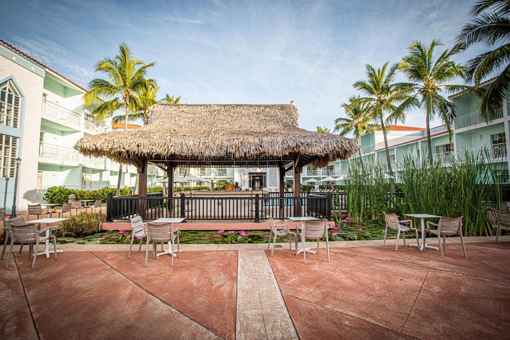 Tropu stila paviljons ar salmu jumtu, ap kuru izvietoti galdi un krēsli, ieskauts zaļumiem un palmu kokiem. Fons veido mierīgu viesnīcas iekšējo pagalmu ar ūdeni un ziediem.