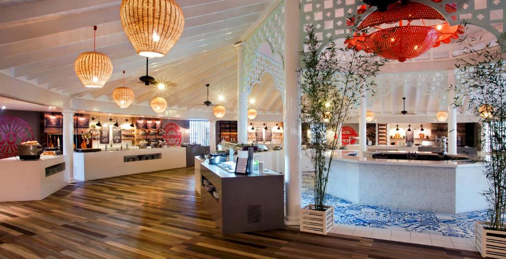 Mūsdienīga restorāna interjers ar koka grīdu, elegantām piekārtiem gaismas ķermeņiem un dekoratīvām augu kompozīcijām. Gaisotne ir silta un viesmīlīga, izceļot ēdināšanas zonu ar stilīgu dizainu.