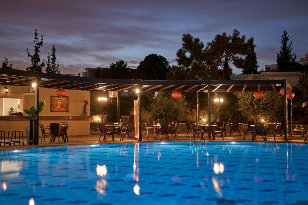 Vakara ainava pie peldbaseina, apgaismota ar siltām gaismām un sēdvietām zem nojumes, radot relaksējošu atmosfēru. Vides elementi, piemēram, augi un mākslas darbi, papildina šarmu un mierīgumu.