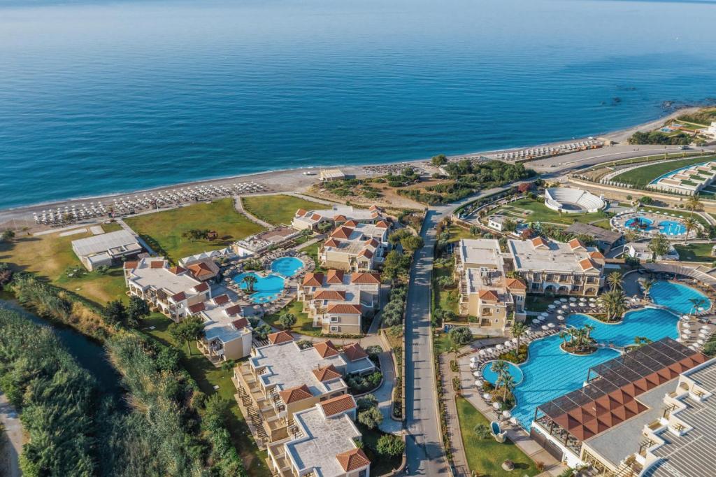 Viesnīcas komplekss ar peldbaseiniem un tuvumā esošo pludmali, kas atrodas pie zilo jūras ūdeņu. Apkārtne ir zaļa ar augiem un ainavām, radot relaksējošu atmosfēru.