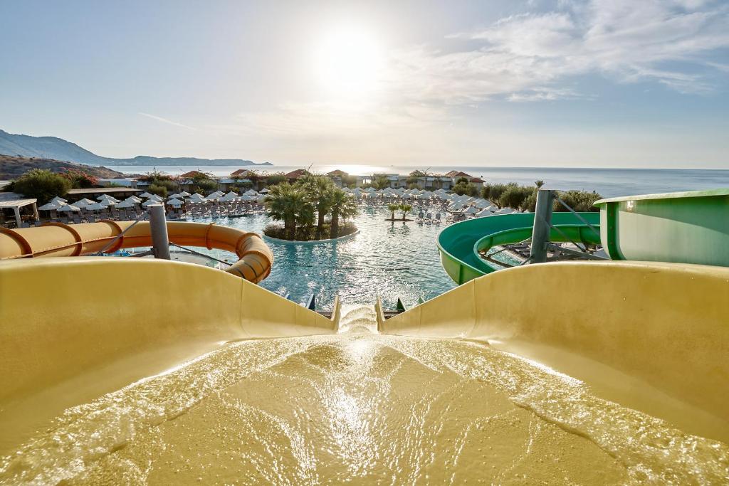 Ūdens slidkalniņš ar skatu uz baseinu un jūru saulainā dienā, apņemts ar palmām un sauļošanās krēsliem. Ideāla vieta atpūtai un izklaidei.