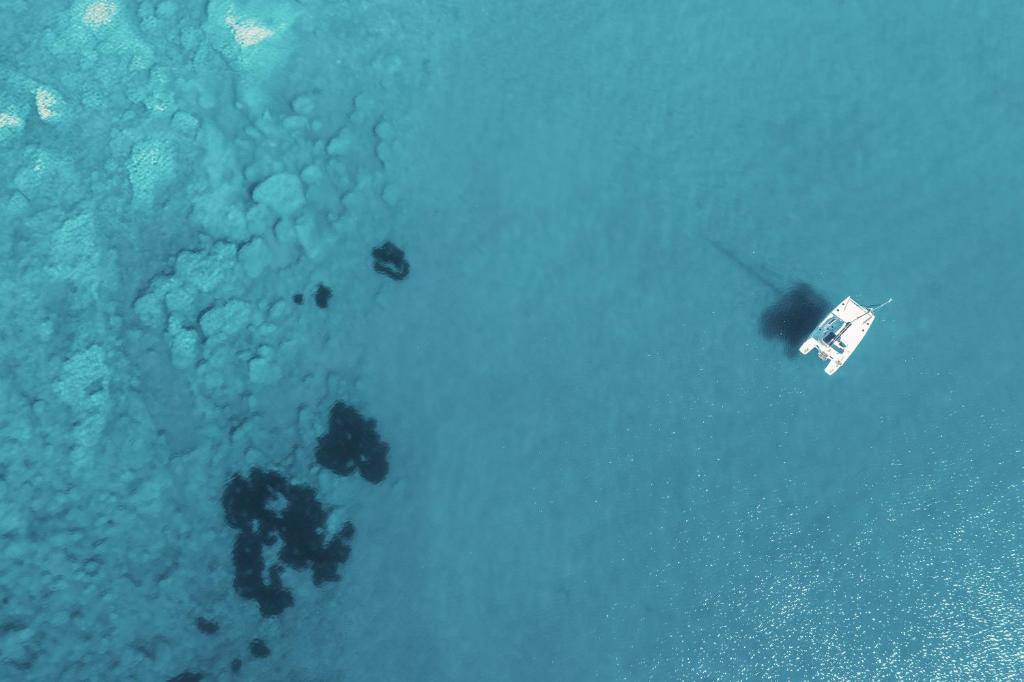 Zilā ūdens virsma ar laivu, kas peld pa vidu. Apakšā redzama jūras augsne un akmeņi.