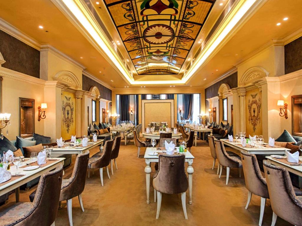 Elegants restorāns ar greznām mēbelēm un dekoratīvu griestu dizainu, kas rada patīkamu atmosfēru. Galdi ir sakārtoti ar izsmalcinātu galda piederumu izvēli un ziediem.