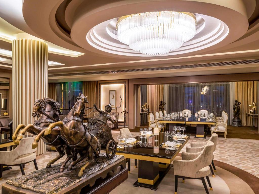 Elegants restorāna interjers ar greznu lustru un bronzas skulptūru, kas attēlo divus zirgus un ratos sēdošu figūru. Galdi ir saklāti ar galda piederumiem, radot izsmalcinātu atmosfēru.
