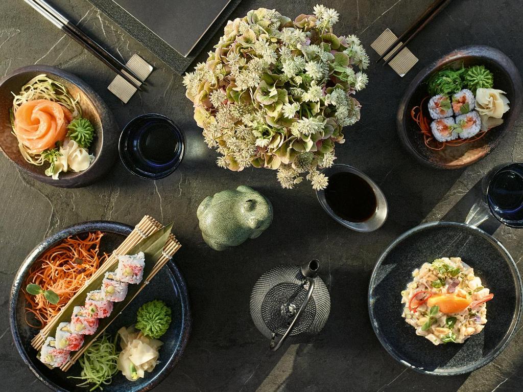 Sushi un japāņu ēdieni izkārtoti uz eleganta melna galda, papildināti ar ziediem un tējas komplektu. Krāsainas sastāvdaļas un rūpīgi sakārtotas šķīvji rada pievilcīgu vizuālo noformējumu.
