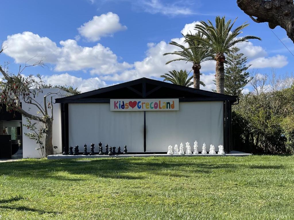 Bērnu izklaides zona ar lieliem šaha figūriem un uzrakstu "Kids❤️Grecoland" virs ieejas, ko ieskauj zaļa zāle un palmas.