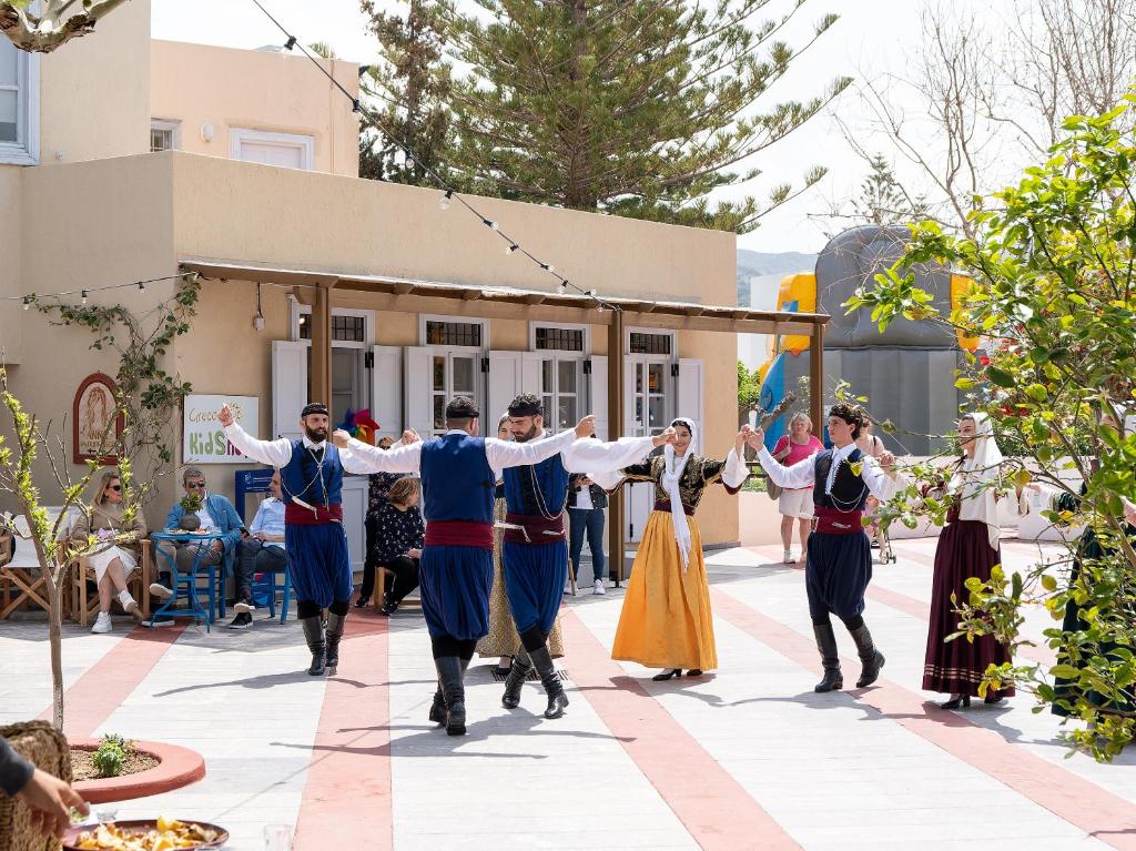 Tradicionāls grieķu tautas dejas priekšnesums ar dalībniekiem tradicionālajos apģērbos, notiek saulainā laukumā. Fons piepildīts ar skatītājiem un zaļām augu kompozīcijām.