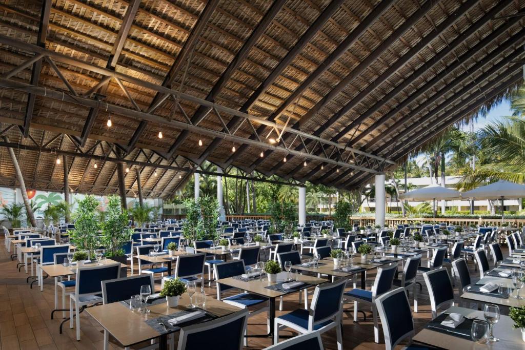 Mūsdienīga restorāna iekštelpa ar plašu zāli, kurā ir elegantas galdi un krēsli, kā arī zaļumi dekorēšanai. Dabas gaisma un tropu ainava veido patīkamu atmosfēru.