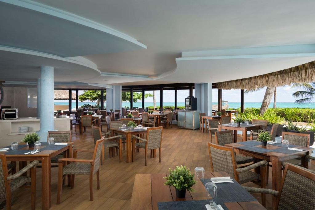 Moderns restorāns ar skatu uz jūru, kur koka galdi un krēsli ir izkārtoti ērtā interjerā. Gaisotne ir relaksējoša ar zaļumiem un dabiskiem elementiem, kas piešķir vietai viesmīlību.