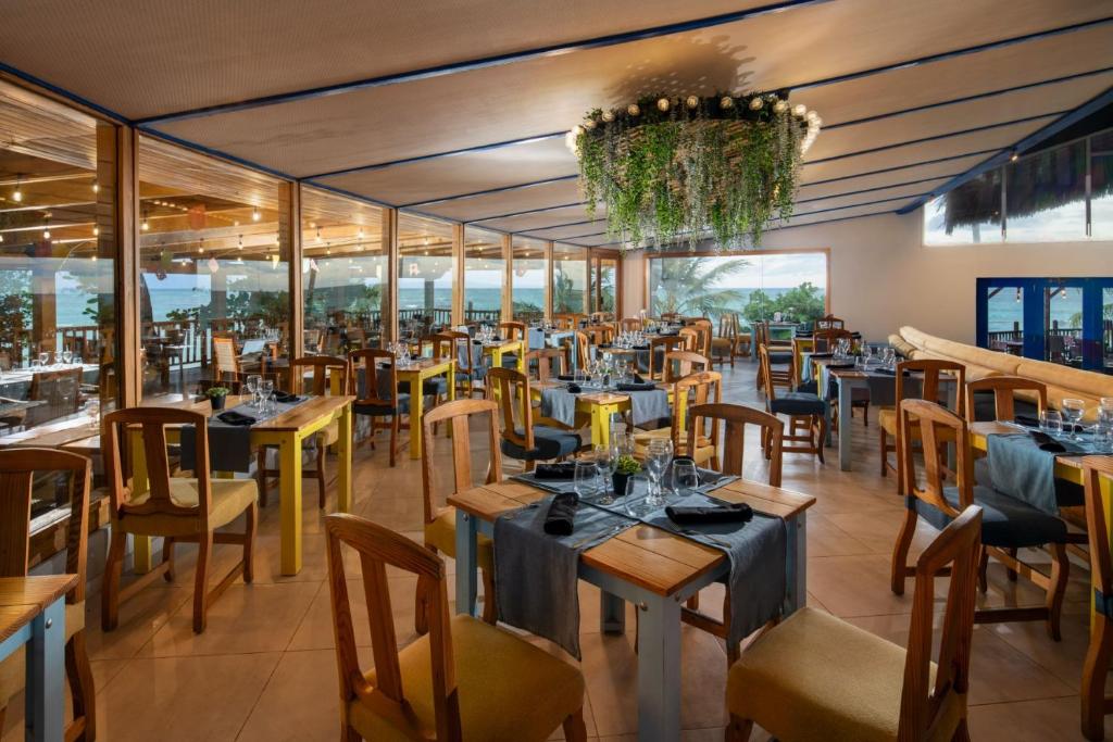 Mūsdienīga restorāna iekārtojums ar koka galdiem un krēsliem, eleganti klāti galdi un plašas logu sienas, kas nodrošina skatu uz dabu. Interjerā ir zaļumi un mājīga atmosfēra, kas aicina apmeklētājus.