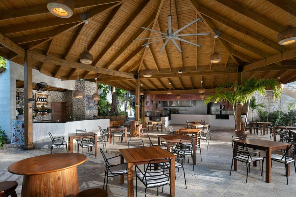 Atvērtas terases restorāna interjers ar koka jumtu, elegantām galdiem un krēsliem, kā arī atvērtu virtuvi. Dabiska gaisma un zaļumi piešķir atmosfēru.