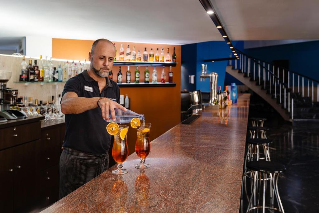 Bāra darbinieks ielej dzērienus glāzēs, rotājot tos ar apelsīnu šķēlēm. Fons ir moderns bārs ar plašu alkoholisko dzērienu klāstu.