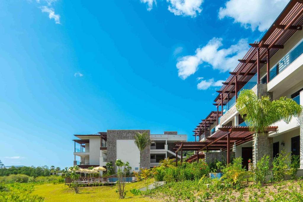 Mūsdienīga viesnīca ar terasēm un zaļo apstādījumu fonu, zem skaidras zila debess.