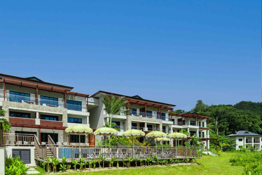 Mūsdienīga kūrorta ēka ar terasēm un dārzu, kurā ir saules parasoles, ieskauta zaļajā dabu. Skats uz skaidru zilo debesu fonu.