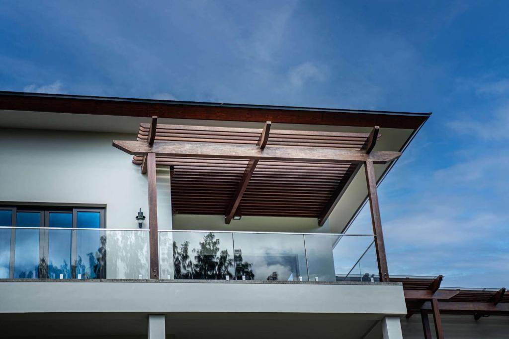 Mūsdienīgas mājas arhitektūra ar koka pergolu un stikla balkonu, kas izceļas pret zilu debesu fona.