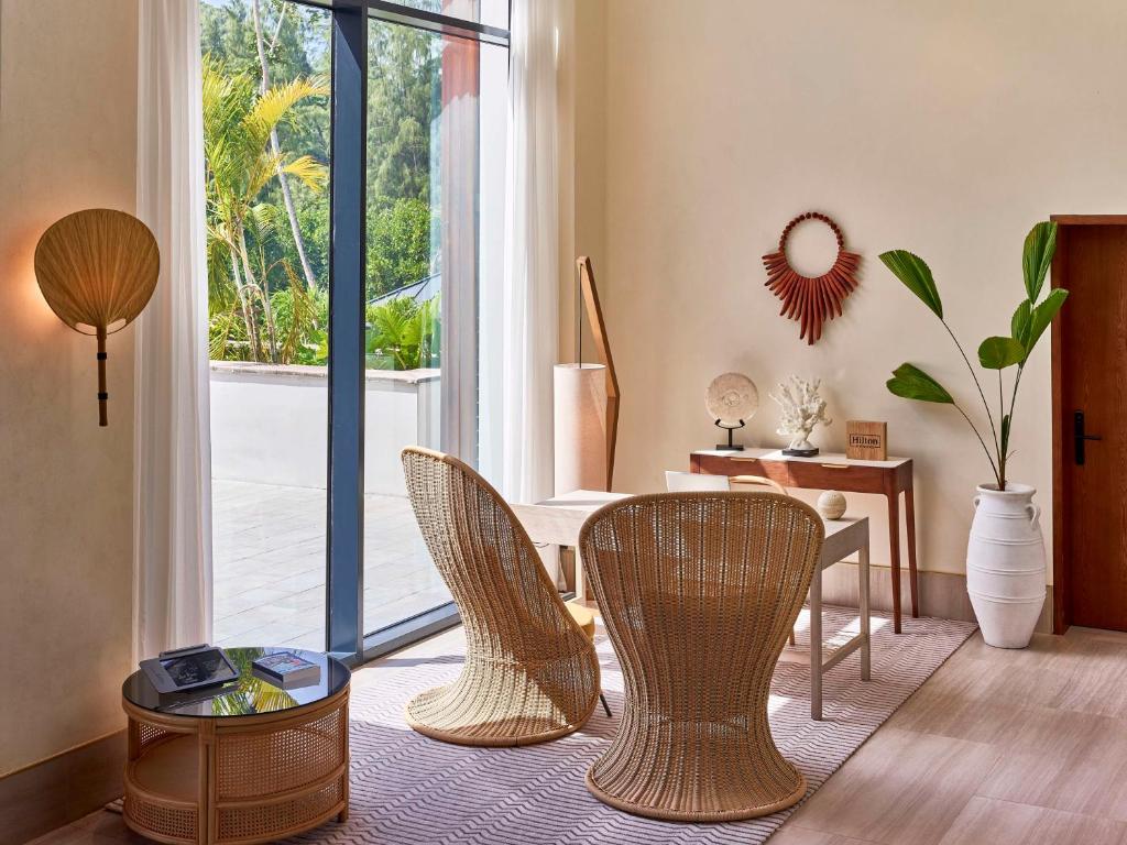 Mūsdienīgs interjers ar rattan krēsliem, koka galdu un daudziem augiem, kas redzami caur lieliem logiem. Gaismas un dabas elementi rada patīkamu un atsvaidzinošu atmosfēru.