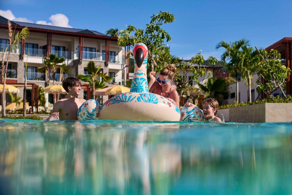 Bērni un pieaugušie izbauda laiku baseinā, spēlējot ar rozā pīli. Apkārt ir tropiskas augu ainavas un viesnīcas ēkas.