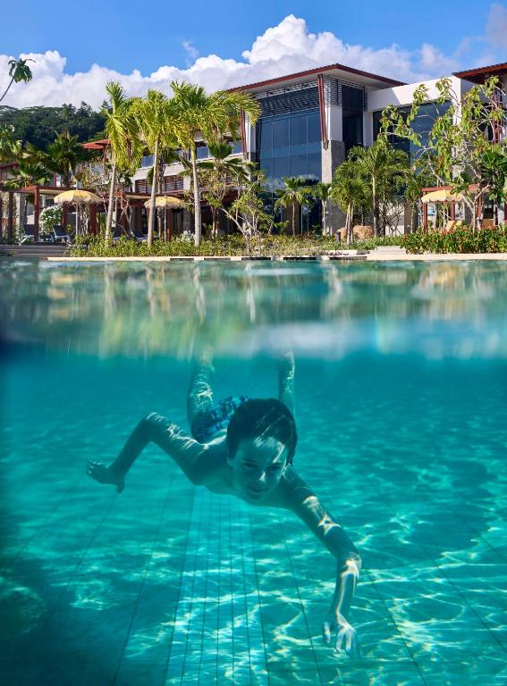Bērns peld zem ūdens skaidrā baseinā, ar fonā redzamu modernu viesnīcu un tropisko ainavu.