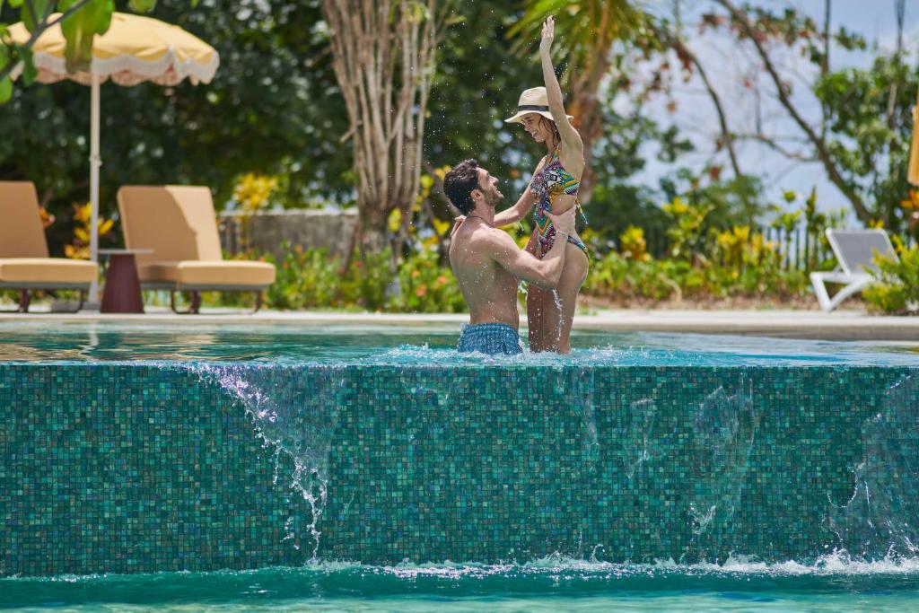 Pāris izbauda laiku pie baseina, kur vīrietis pacēlis sievieti gaisā, radot jautru un romantisku atmosfēru. Apkārt ir zaļumi un sauļošanās krēsli.