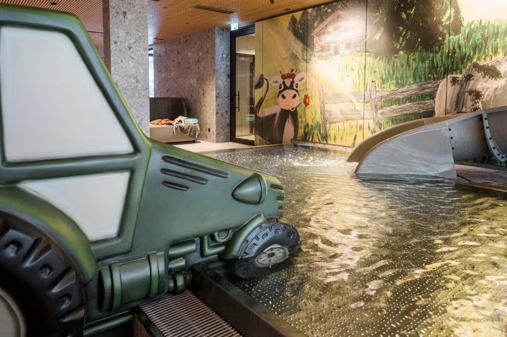 Bērnu ūdens spēļu zona ar zaļu traktoru un krāsainu grafiti fona, kur attēlota govs un lauku ainava.